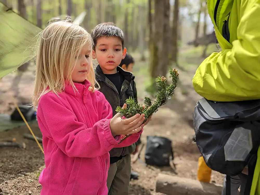 A student eagerly presents a branch to the facilitator for inspection, while a friend watches attentively. Both the student and the friend display curiosity and anticipation, engaging in a moment of learning and exploration in the natural environment.