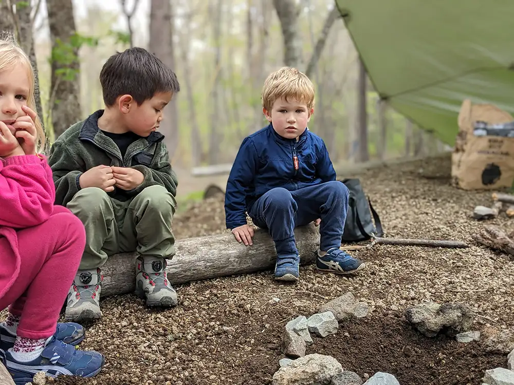 Friends gather in a circle around the pit they have prepared for the campfire, eagerly awaiting the next step. Their faces filled with anticipation and excitement. The pit is ready, and soon they will add the sticks they collected to fuel the fire.