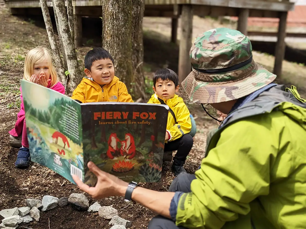 
Head Camp Facilitator, Kai Chang, reads "Fiery Fox" to students at Rising Field in Karuizawa, teaching them about fire safety. Students listen attentively, engrossed in the interactive storytelling experience, while learning important lessons in a serene forest setting.